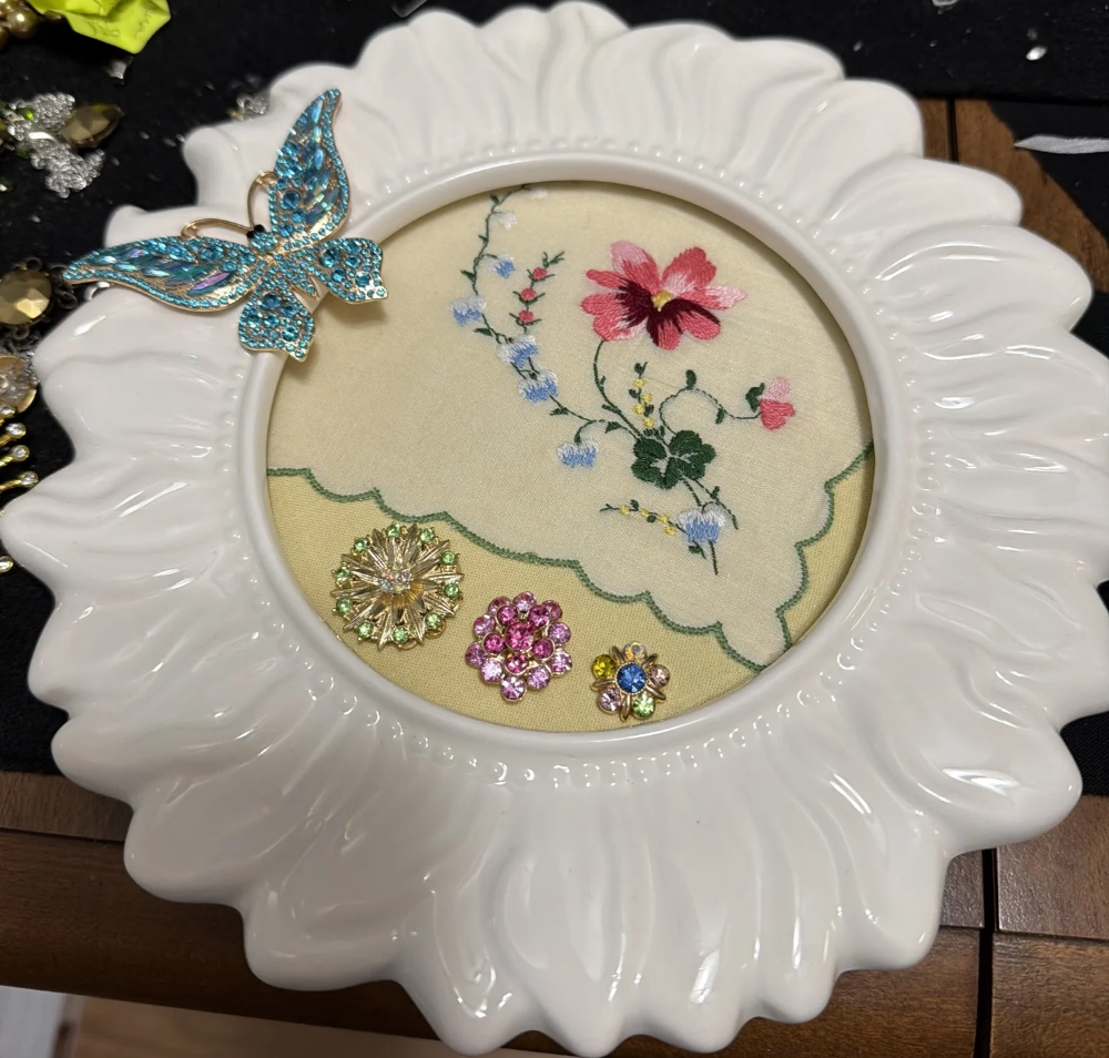 A round white ceramic frame shaped like a sunflower displaying embroidered fabric with vintage brooches inside.
