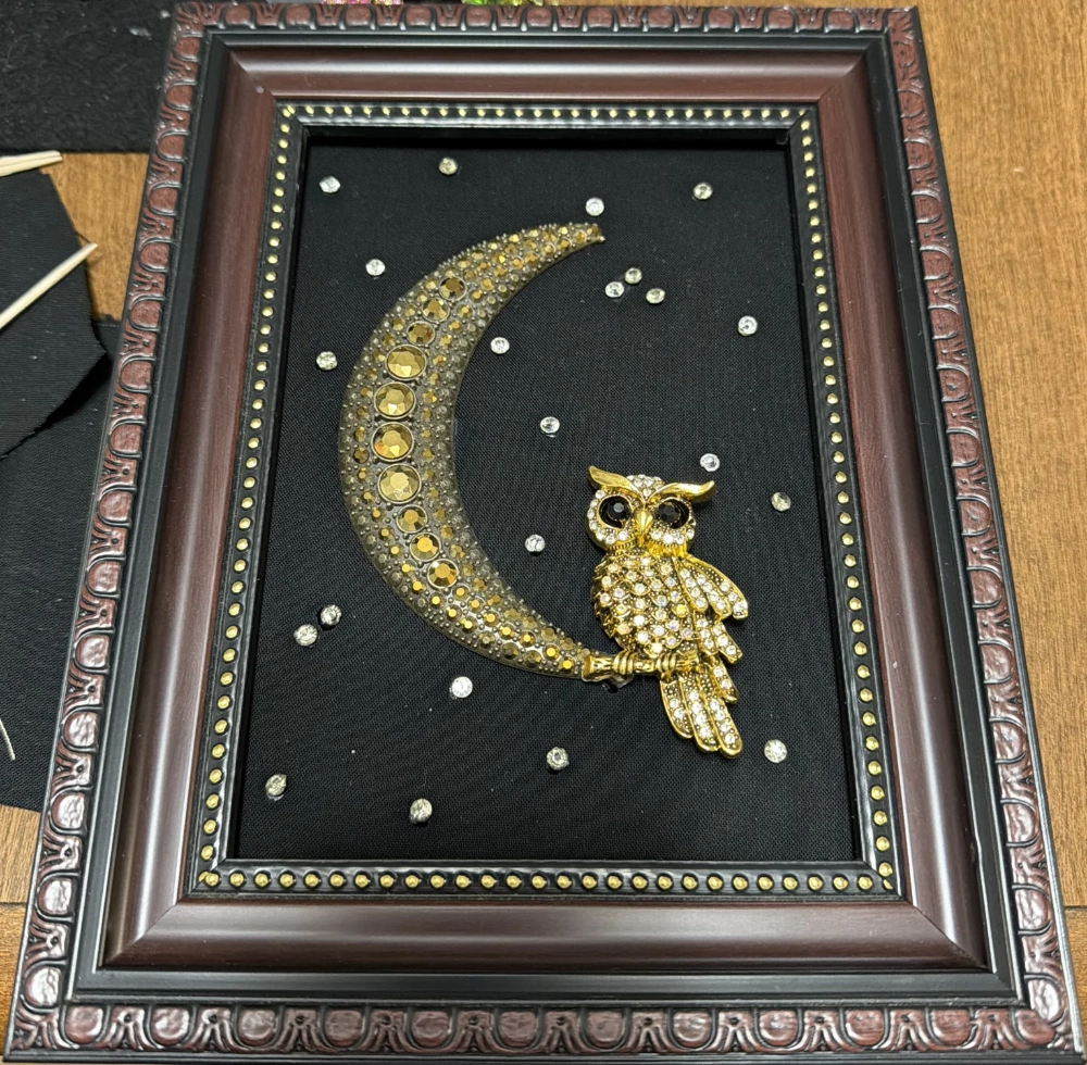 An ornate dark wood frame with gold inner trim displaying a gold rhinestone owl sitting on a textured gold crescent moon against a black background with scattered crystals.