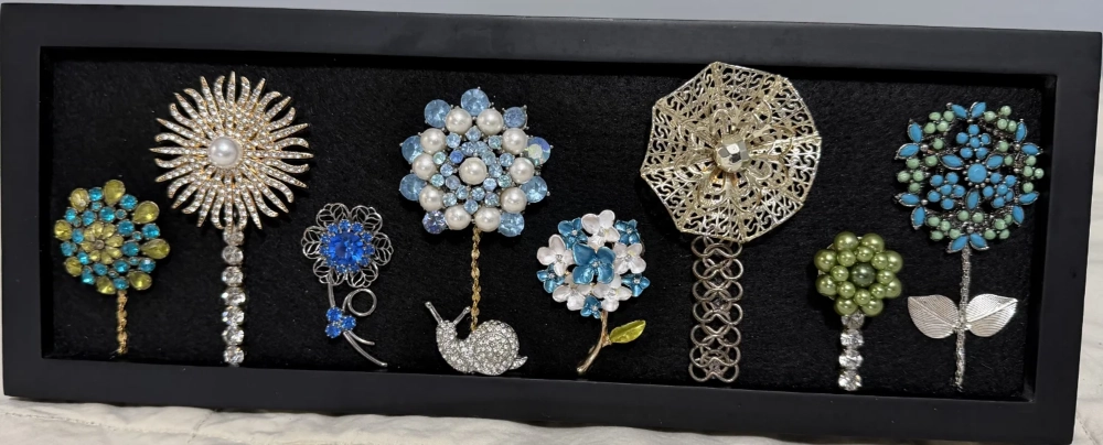 A long rectangular black frame displaying a row of eight unique vintage rhinestone flower brooches arranged like a garden.