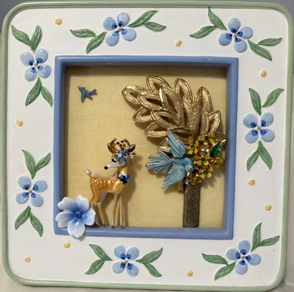 A square ceramic frame with sculpted blue flowers enclosing a vintage enamel deer and a blue bird perched by a golden metallic tree.