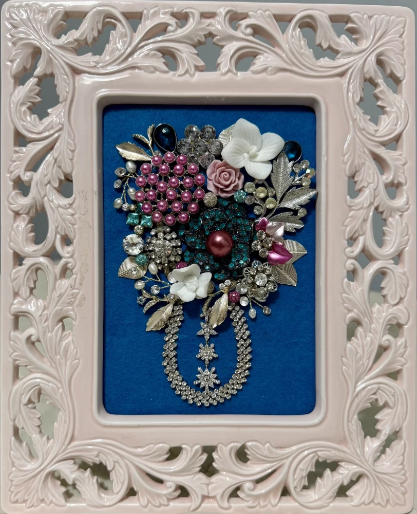 An ornate light pink baroque frame enclosing a rich bouquet arrangement of vintage brooches and rhinestone strands on a solid blue backdrop.