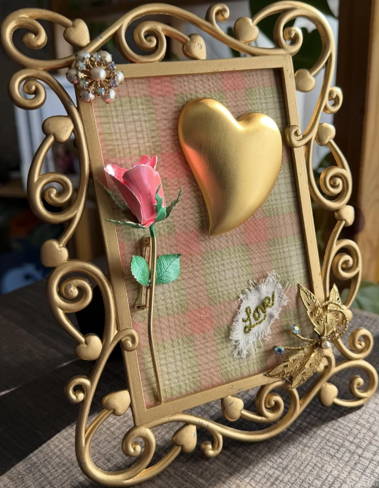 An ornate gold open-scrollwork frame showcasing a large matte gold heart, a pink enamel rose brooch, and an embroidered "Love" patch on plaid fabric.