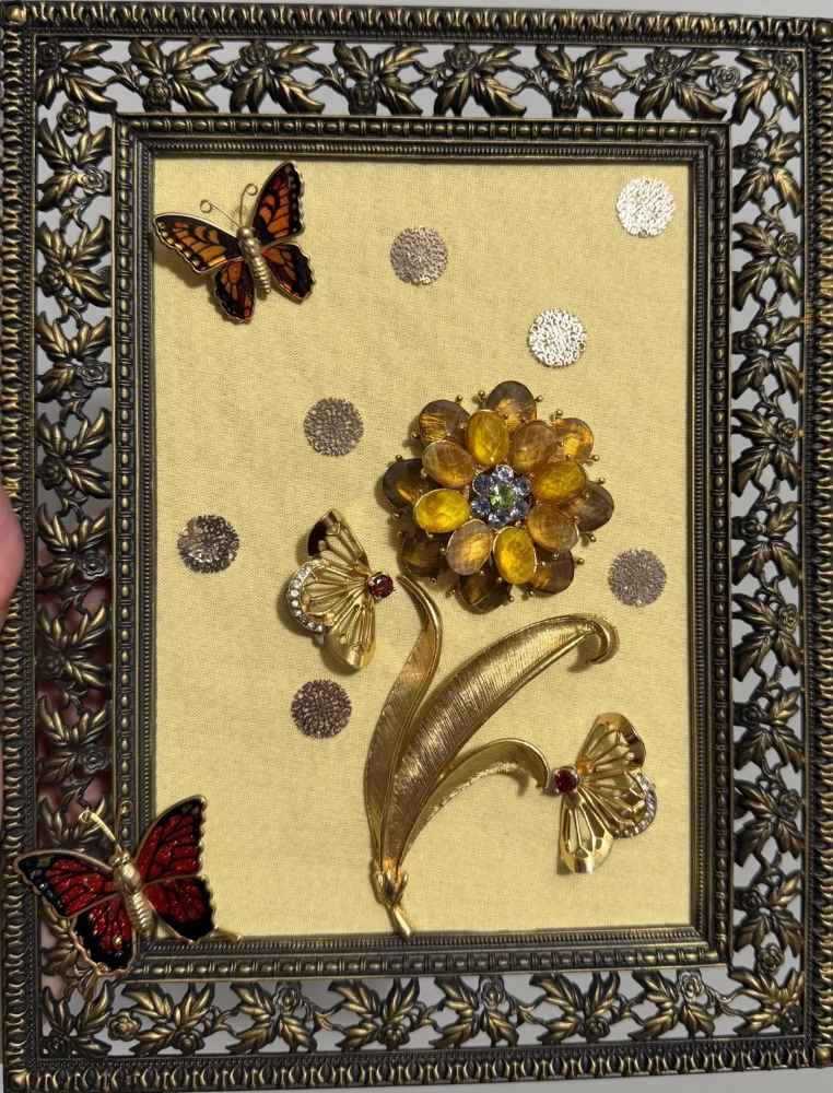 An ornate bronze openwork frame enclosing a large amber rhinestone flower brooch and multiple vintage butterfly pins on a pale yellow fabric backing.